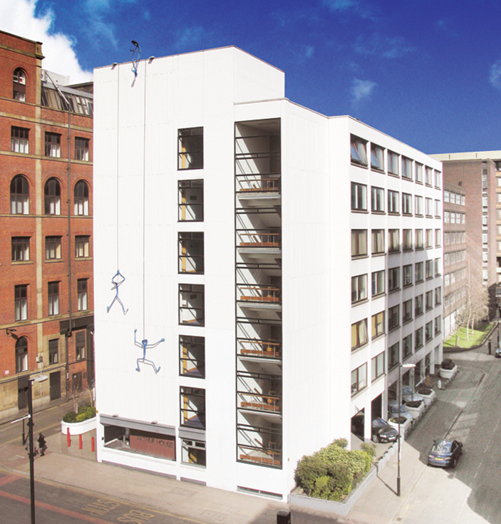 Auf einer weißen, mehrstöckigen englischen Schule sind Strichmännchen zu sehen, die so aufgemalt sind, als würden sie die Außenwand des Treppenhauses hinunterklettern, wobei die umliegenden städtischen Gebäude sichtbar sind.